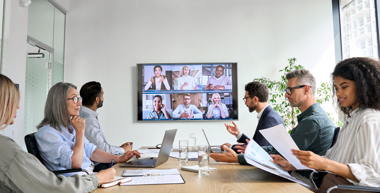 Hybrid meeting taking place in a physical office space with participants on a television who have used videoconferencing to join the meeting