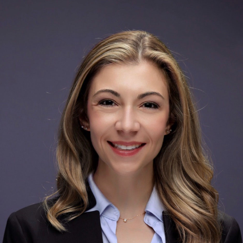A professional headshot of a woman with long, wavy blonde hair and a friendly smile, wearing a black blazer over a light blue collared shirt and a delicate gold necklace against a solid grey background.