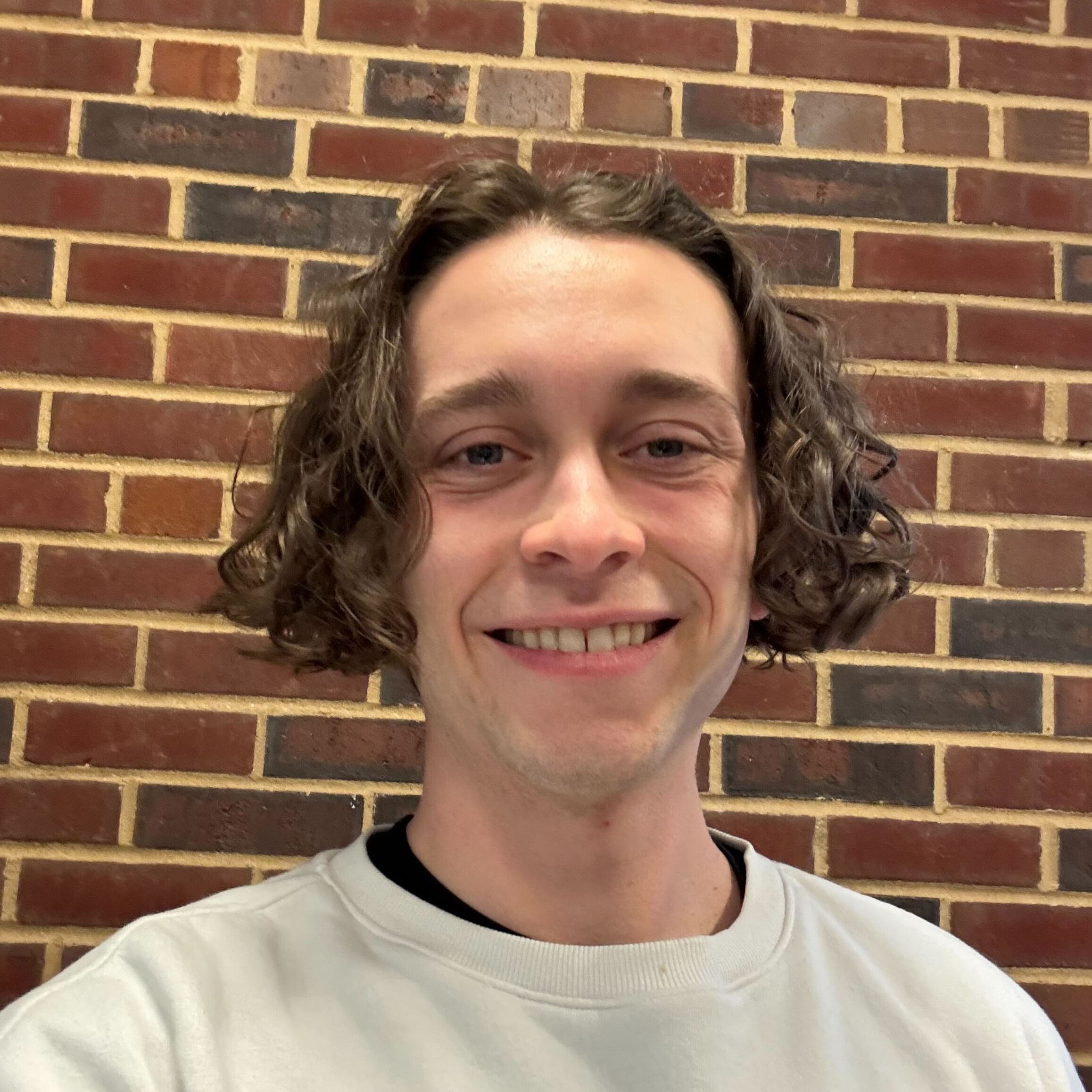A young man with brown curly hair and a white sweatshirt smiling in front of a multicolored brick wall.