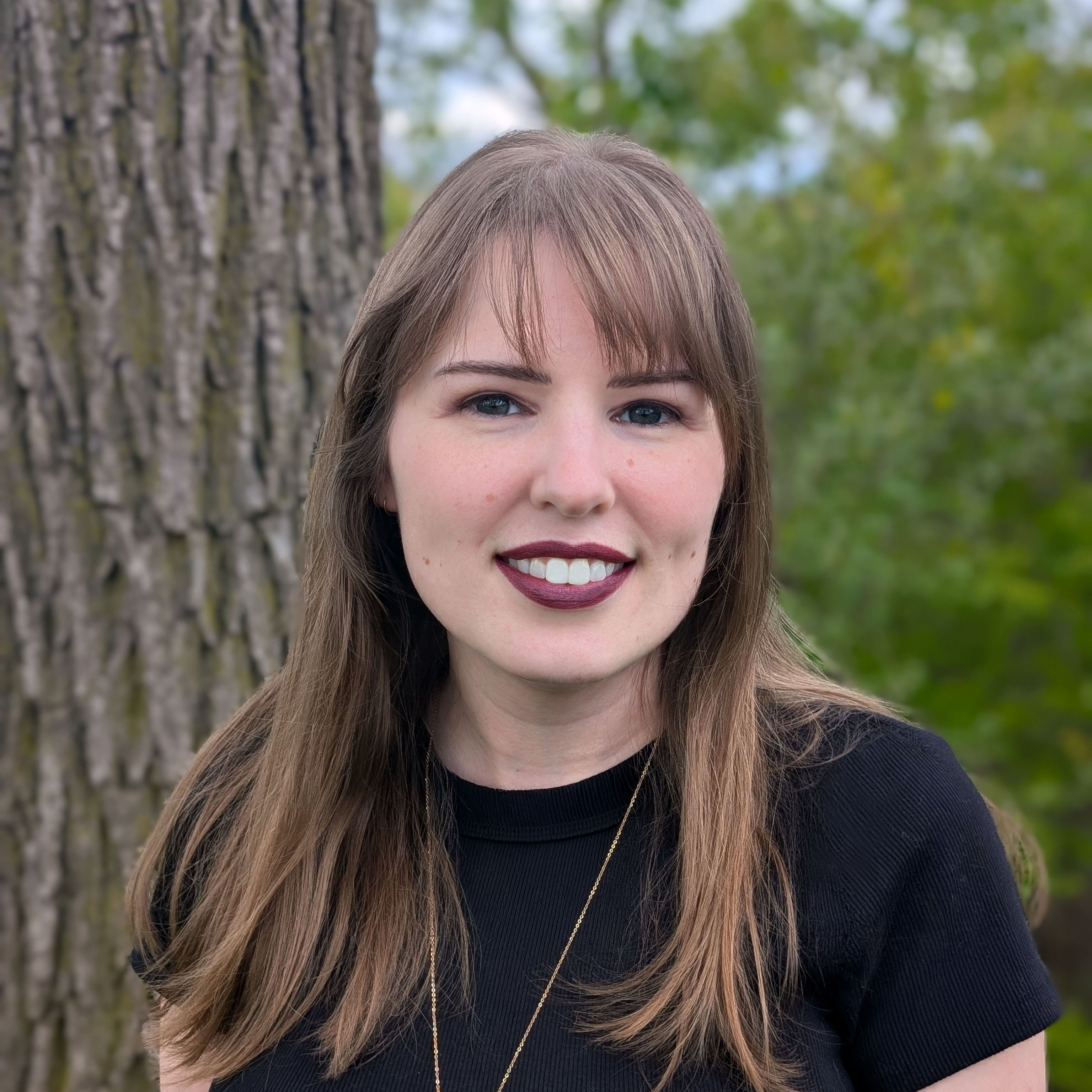 A headshot of a woman with long light brown hair and bangs, smiling in front of a blurred outdoor background of trees. She is wearing a black shirt, a thin gold necklace, and dark berry-colored lipstick.