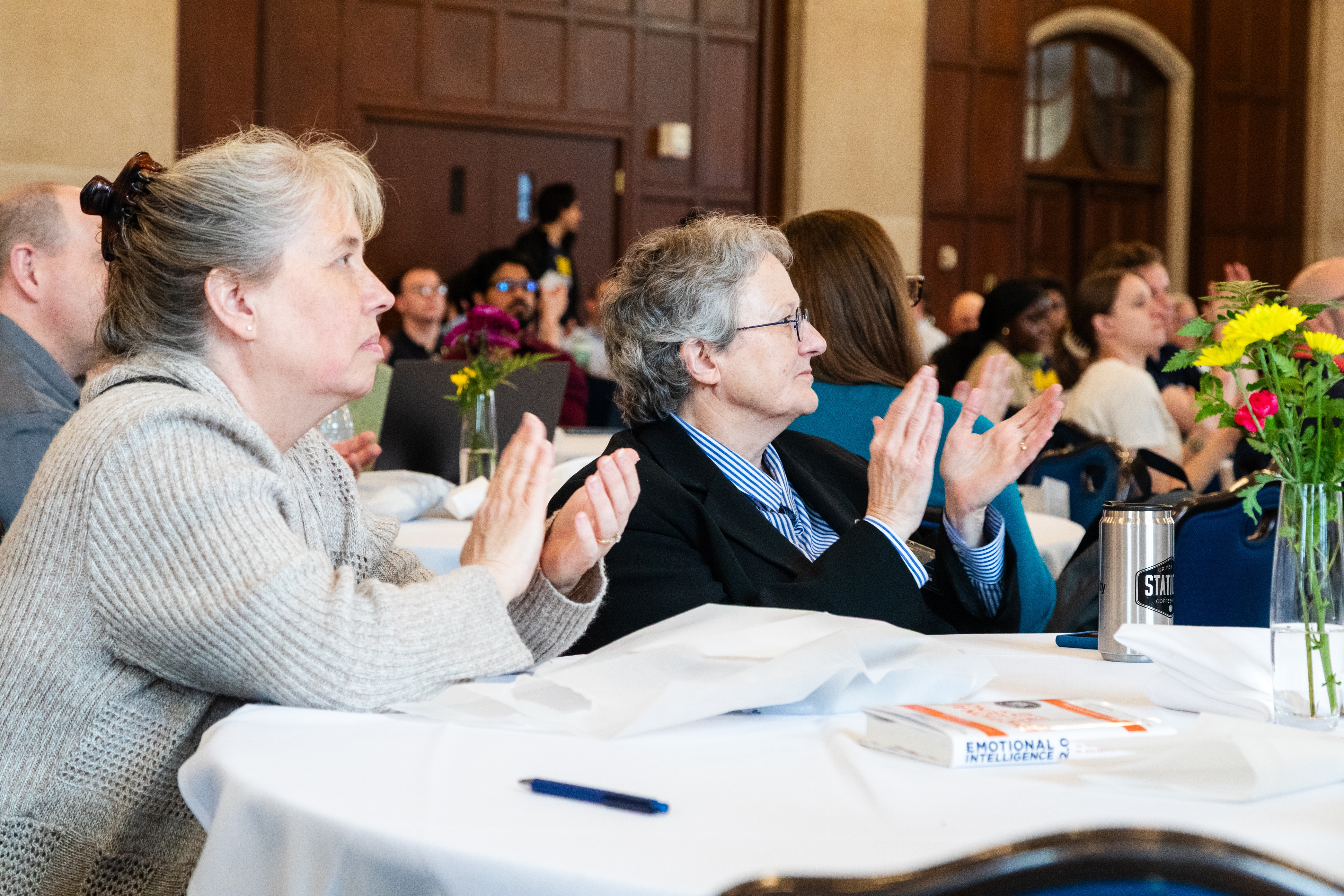 Two attendees sit in profile, looking up at the stage. They are both clapping.