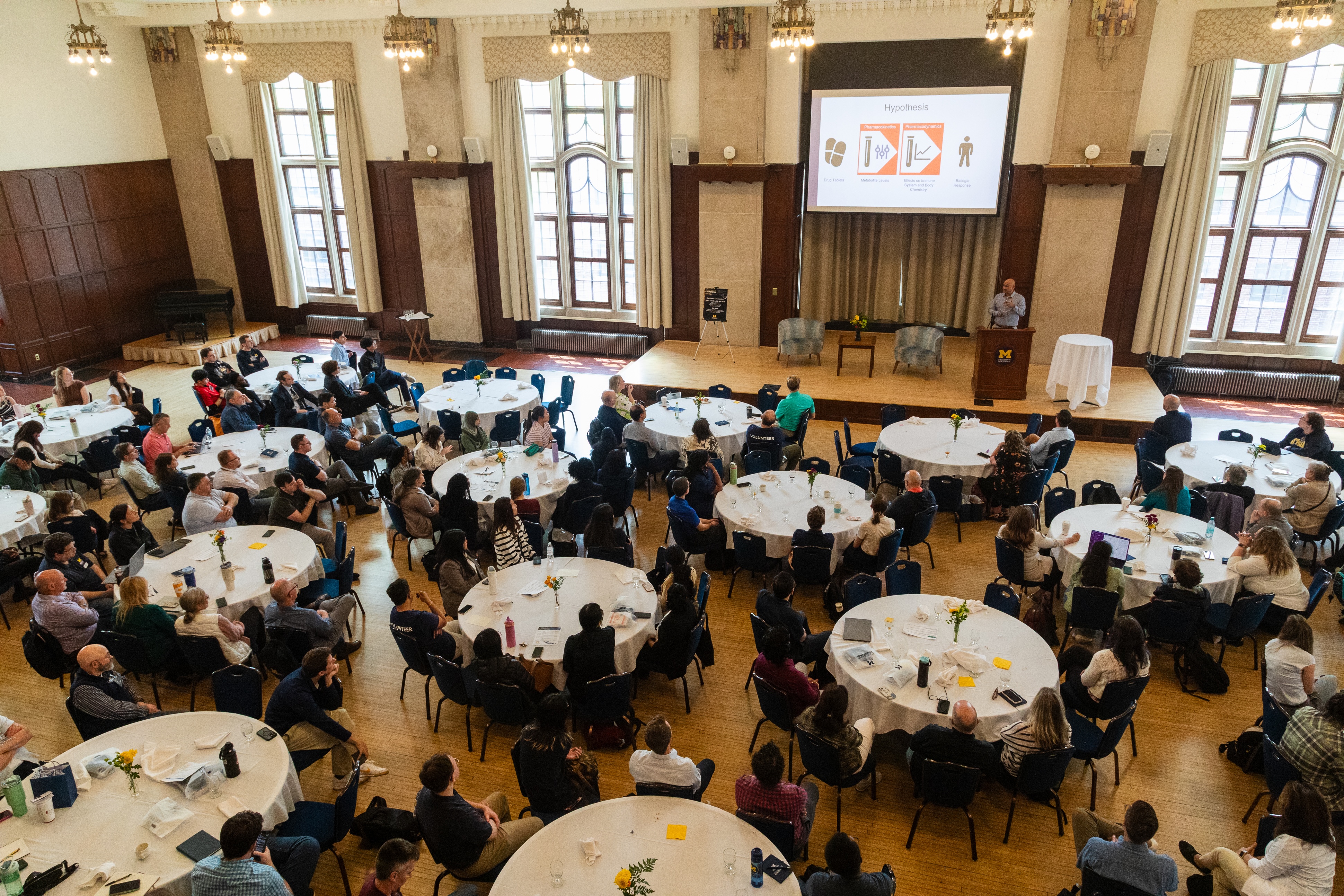 An aerial photo of a ballroom filled with attendees sitting at round tables. A presenter stands at a podium on stage.