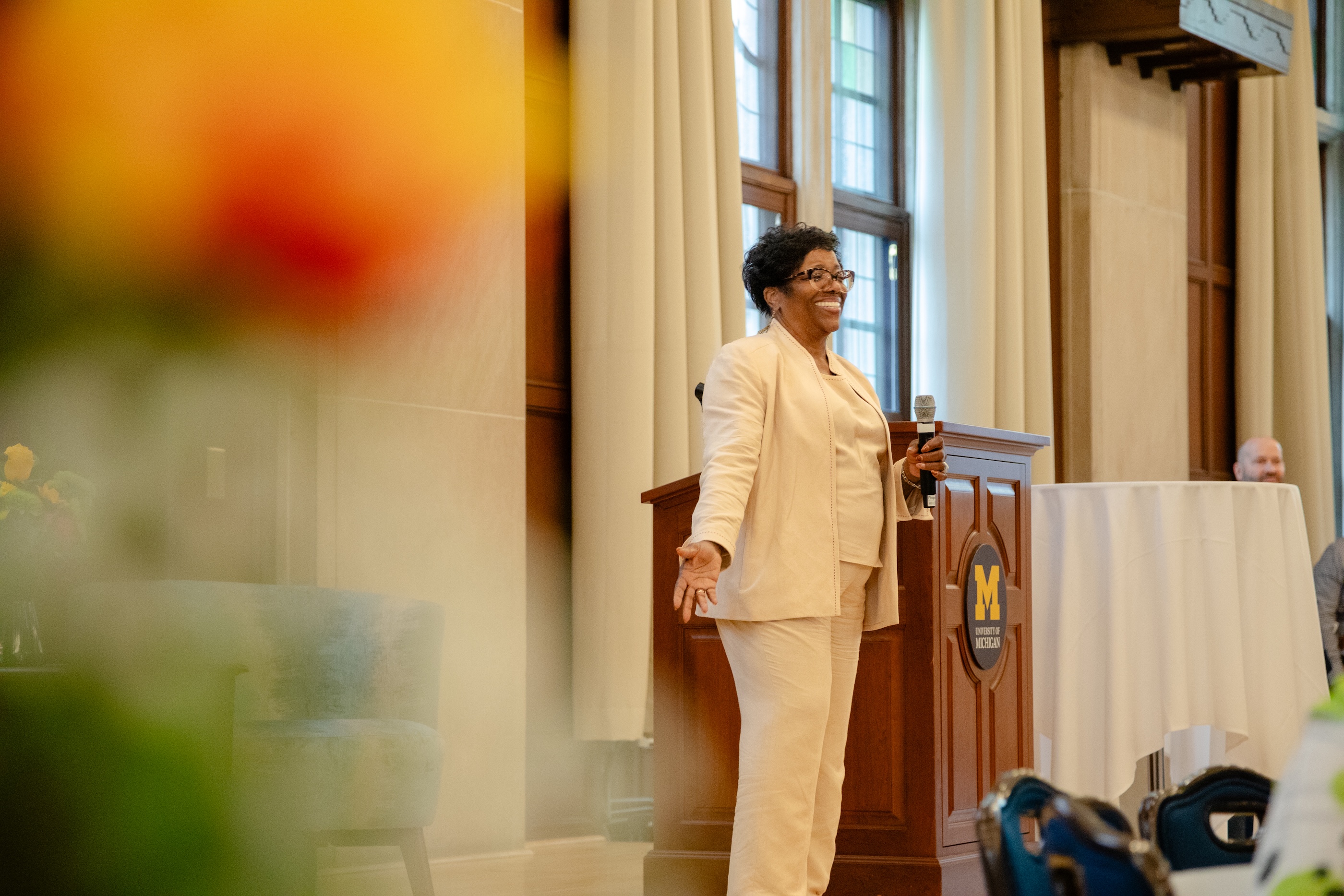Diane Jones stands on stage, smiling, while holding a microphone. She is standing next to a U-M podium.